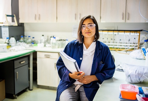 Portrait of a confident young research scientist wearing proctective glasses sitting at her desk surrounded by laboratory equipment