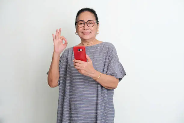 Elderly Asian woman smiling confident and give OK sign while holding mobile phone