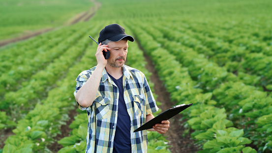 Male farmer in agricultural field holding a clipboard and talking on walkie-talkie