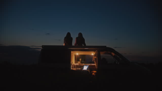 Dolly shot of a silhouette of two women relaxes on the roof of a camper parked in the field at night. An open laptop in the cabin of a camper. Shoot in 8K resolution.