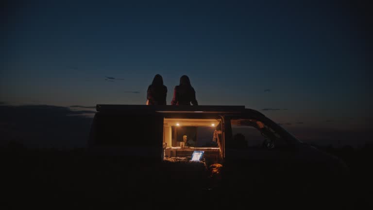 DS Two women relaxes on the roof of a camper in the middle of a field at night