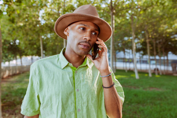 Portrait of young cheerful stylish multiracial homosexual man talking with smartphone at park. Wears green shirt and hat outdoors.