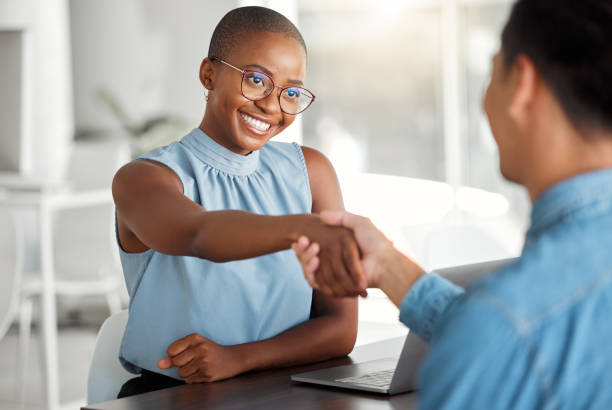 dois empresários alegres apertando as mãos em uma entrevista juntos no trabalho. colegas felizes cumprimentando com um aperto de mão em um escritório. chefe afro-americana promovendo um empregado do sexo masculino - gesturing interview business sitting - fotografias e filmes do acervo