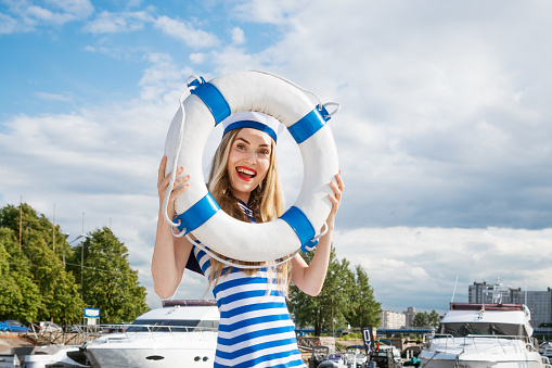 Young happy woman of Caucasian appearance in a blue striped dress standing on a yacht posing with a lifebuoy in her hand, against the background of a blue sky with clouds on a summer sunny day