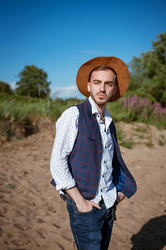 Young handsome man in a shirt and a hat on nature looks to the side during the day in summer