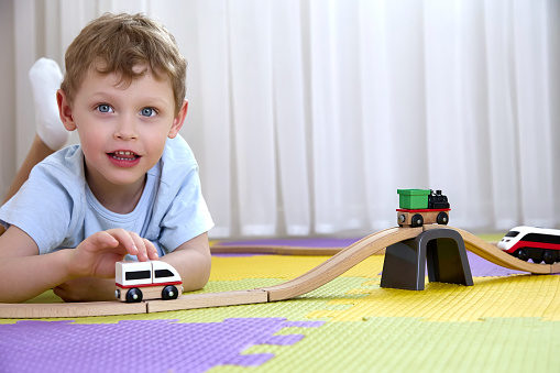 Funny boy 4-5 years old looks into the camera and plays with a train and a wooden railway. A child plays with toys on puzzle mats in the playroom.