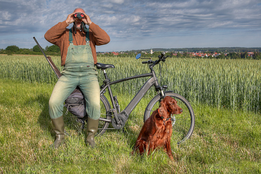Hunter sits on the luggage rack of his bike and watches the hunting area in front of the small town through his binoculars. Next to him in the grass sits his Irish Setter hunting dog.