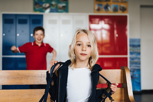 portrait of elementary school boy sitting on bench in school corridor and looking at camera, other one fooling around behind him