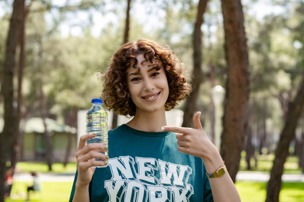 Happy redhead woman wearing green sportive clothes standing on city park, outdoors drinking bottle of water for refreshment very happy pointing with hand and finger. stock photo