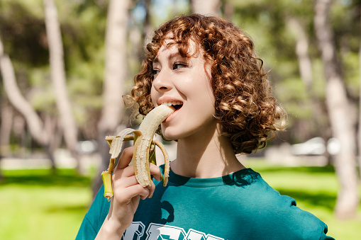 Happy young sportive woman wearing green t-shirt standing on city park, outdoors biting healthy banana for strength and energy. Outdoor sports and healthy food and life concepts.