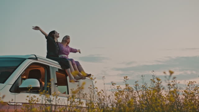 WS Two women in embrace sit on the roof of a camper parked in the field of canola at dusk
