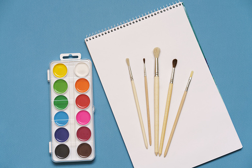 a set of brushes, a sketchbook, and bright watercolors on a blue background. the concept of creativity