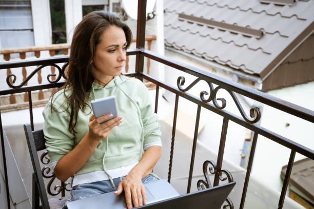 istock Young woman sits on a balcony at a table working with a phone behind a laptop 1400678467