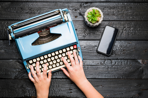 Top view of mans hands using vintage blue typewriter placed on dark wooden desk