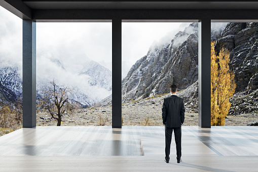 Thoughtful young european man standing in bright modern concrete terrace or balcony interior with beautiful nature view and bright sky. Architecture and design concept