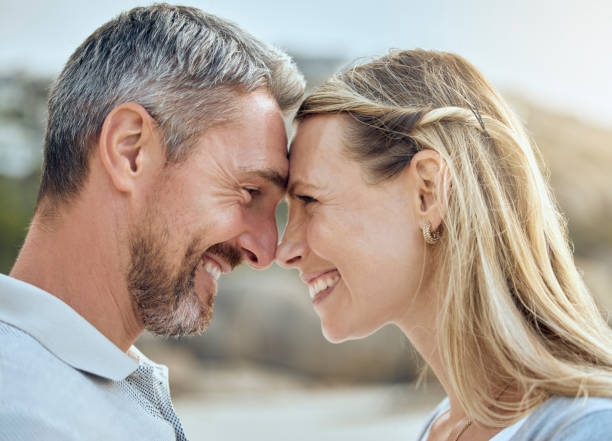 casal caucasiano feliz e amoroso curtindo um encontro romântico na praia juntos em um dia ensolarado. marido e mulher alegremente carinhosos de pé cara a cara com narizes tocando para beijo esquimó enquanto se conectam nas férias ao ar livre - rubbing noses - fotografias e filmes do acervo