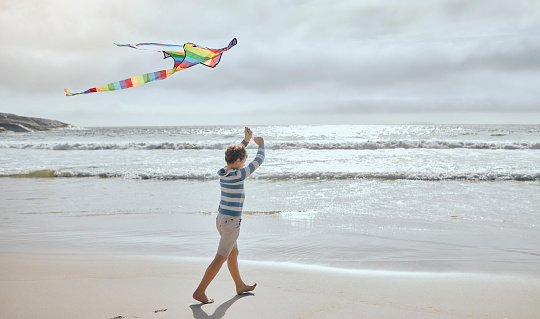 One little caucasian boy flying a colourful rainbow kite in the wind at the beach. Playful young child having fun outdoors. The innocence of childhood