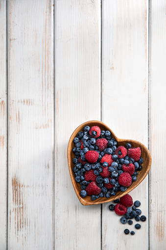 This is an overhead photo of a heart bowl of berries on a white wood background