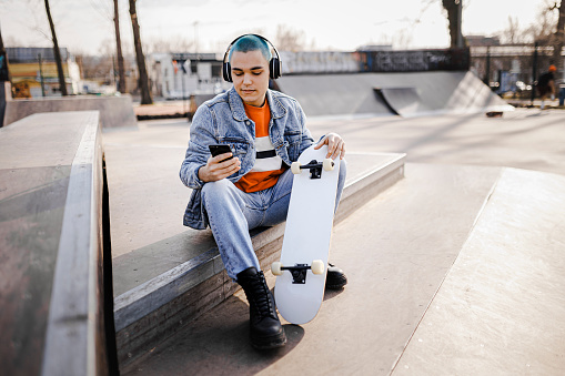 Boy  relaxing with his skateboard listening to music. Urban boy enjoying listening to music from her mobile phone while sitting at skatepark.