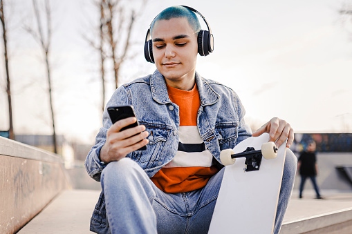 Boy  relaxing with his skateboard listening to music. Urban boy enjoying listening to music from her mobile phone while sitting at skatepark.