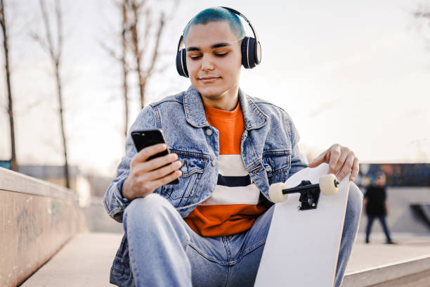 Boy  relaxing with his skateboard listening to music. Urban boy enjoying listening to music from her mobile phone while sitting at skatepark. Boy  relaxing with his skateboard listening to music. Urban boy enjoying listening to music from her mobile phone while sitting at skatepark. Iphone stock pictures, royalty-free photos & images