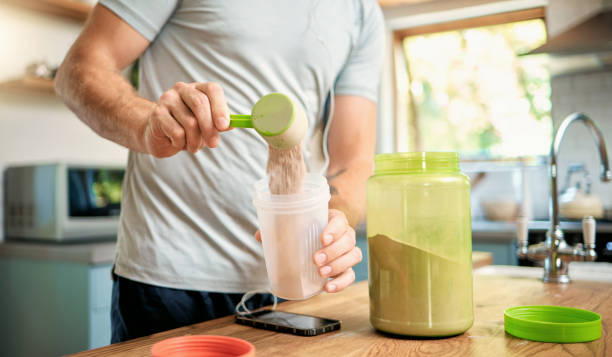 primer plano de un hombre caucásico vertiendo una cucharada de proteína de suero de chocolate en polvo a un batido de salud para obtener energía para entrenar el entrenamiento en una cocina en casa. chico que tiene un suplemento deportivo nutricional pa - protein fotografías e imágenes de stock