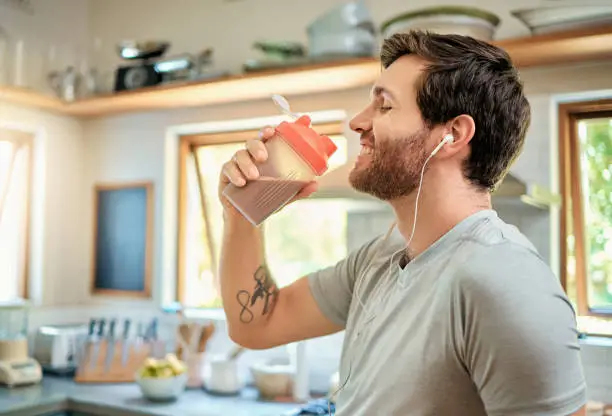 One fit young caucasian man drinking bottle of chocolate whey protein shake for energy for training workout while wearing earphones in a kitchen at home. Guy having sports supplement for muscle gain and dieting with weightloss meal replacement