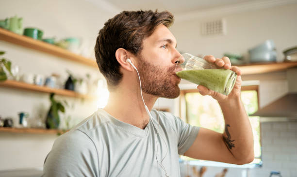 un giovane caucasico in forma che beve un bicchiere di frullato disintossicante verde sano mentre indossa gli auricolari in una cucina di casa. ragazzo che ha succo di frutta fresca per pulire e fornire energia per l'allenamento. bevanda salutare con vitam - juice glass healthy eating healthy lifestyle foto e immagini stock