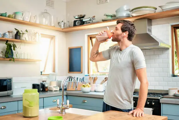 Photo of One fit young caucasian man drinking bottle of chocolate whey protein shake for energy for training workout while wearing earphones in a kitchen at home. Guy having sports supplement for muscle gain and dieting with weightloss meal replacement