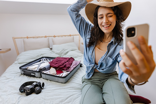 Young woman sitting on the bed, packing suitcase and taking selfie. She is winking.