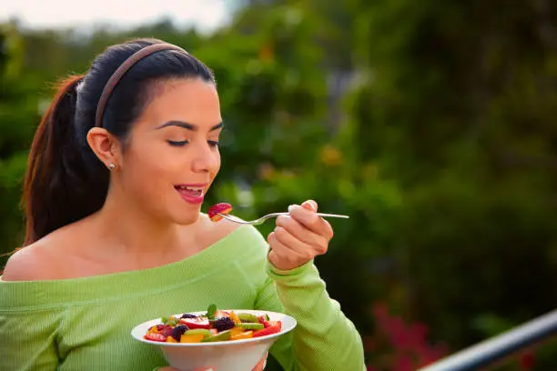 Vegan food themes. Close-up of a Hispanic cute young woman eating fruit salad at lunch outdoors with copy space.