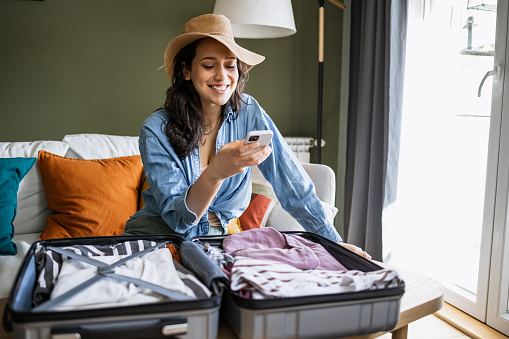 Young woman sitting on the couch, packing suitcase and texting