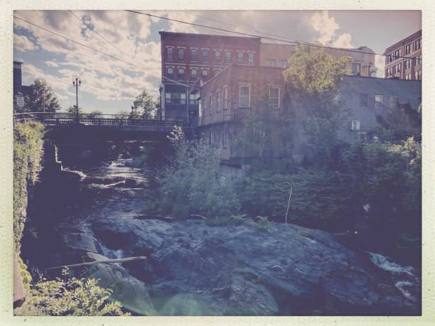 Historic stone mill with a red roof stands near a rocky river. Autumn trees with bare branches and sparse foliage surround the area. A rustic stone bridge crosses the river, which flows calmly through a rural landscape. The scene captures the tranquility of the countryside, with reflections of the trees and sky visible on the water's surface. The architecture and natural elements indicate a blend of heritage and nature.