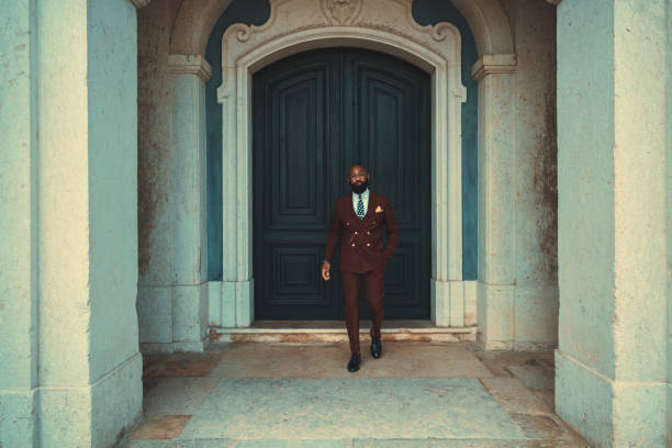 An elegant mature bald bearded African man in a costume and glasses is leaving a wooden entry gate of an antique building; a dapper black guy in a tailored suit walked out the door of an old house