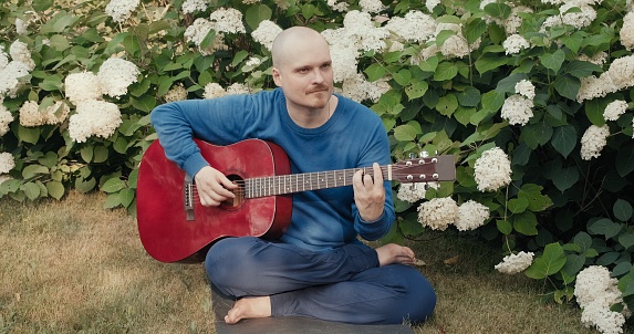 Caucasian man is sitting in a park among flowers and playing a red acoustic guitar. The concept of travel, hobby hikes and lifestyle