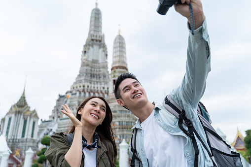 Asian couple backpacker using camera selfie and take a picture in city. Attractive man and woman tourist traveler travel on street, using smartphone record vlog on holiday vacation trip in Thailand.