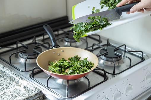 Female hands cooking ora-pro-nobis in frying pan. Cutting board and a knife. Pereskia aculeata is a popular vegetable in parts of Brazil