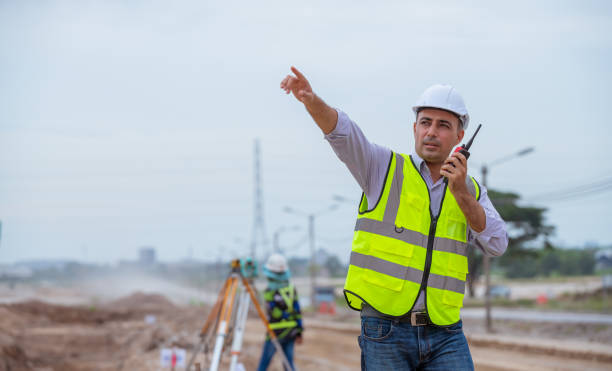 bauingenieur trägt sicherheitsuniform unter inspektion und vermessung arbeitsplatz durch funkkommunikation mit aushubwagen graben, theodolit und arbeiter bau straße hintergrund. - construction safety mid adult men road construction stock-fotos und bilder