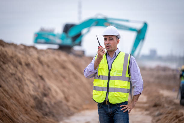 bauingenieur trägt sicherheitsuniform unter inspektion und vermessung arbeitsplatz per funk kommunikation mit bagger lkw graben, theodolit und arbeiter bau straße hintergrund. - construction safety mid adult men road construction stock-fotos und bilder