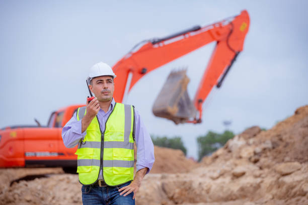 bauingenieur trägt sicherheitsuniform unter inspektion und vermessung arbeitsplatz per funk kommunikation mit bagger lkw graben, theodolit und arbeiter bau straße hintergrund. - construction safety mid adult men road construction stock-fotos und bilder