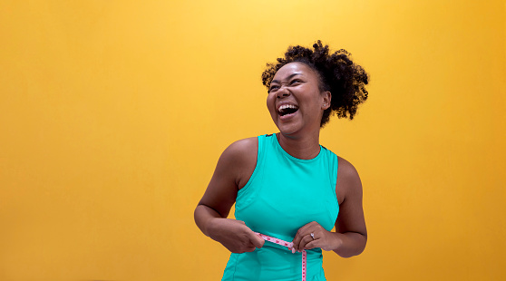 Happy African American woman in sportswear is happy while measuring waist with tape isolated on yellow background for healthy food and exercise