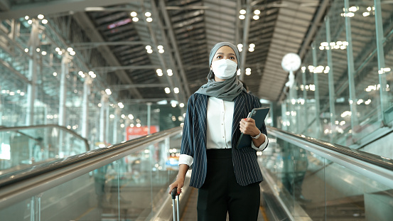 An Asian muslim woman in face mask going on an escalator on her way to check-in with her luggage and mobile tablet.
