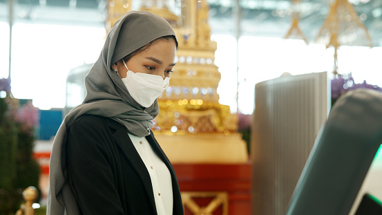 An Asian muslim woman in face mask using a self-service check-in booth provided in the airport.