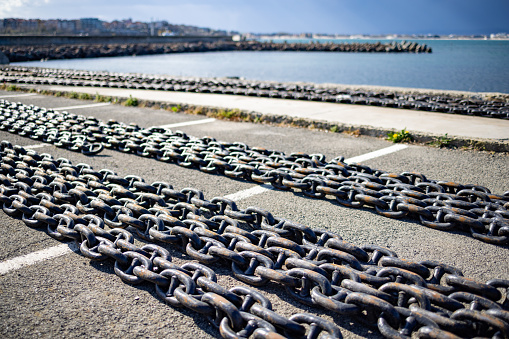 Large long sea chains made of rusty old dark gray metal for ships and boats lie on a wide stone empty pier, against the background of a cloudy blue spacious sky and a quiet calm blue Black Sea