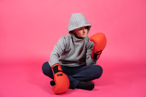 Boxer boy wearing sport clothes sitting on ground isolated on pink. Teenager looking at boxing gloves