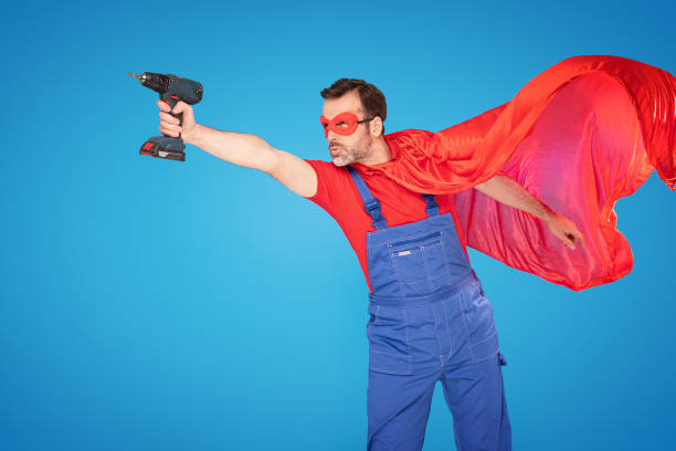 handsome handyman in red cape and mask as a super hero posing with drill in one hand, isolated on blue studio background . repair home concept. - drill red work tool power imagens e fotografias de stock