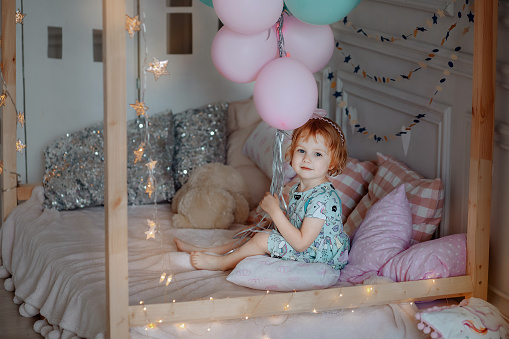 A little child girl in a blue dress sits on the bed and holds blue and pink balloons. Holiday birthday. Didn't like the gift.