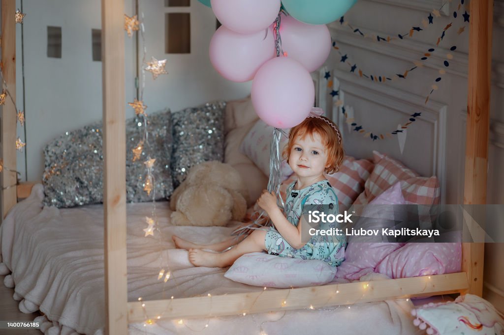 Little baby girl in a blue dress is sitting on the bed. A little child girl in a blue dress sits on the bed and holds blue and pink balloons. Holiday birthday. Didn't like the gift. Girls Stock Photo