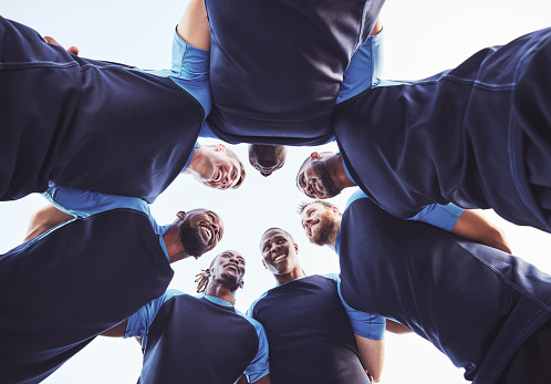 Below diverse group of rugby players standing in a huddle together outside on a field. Young male athletes looking serious and focused while huddled together as a team. Ready for a tough game