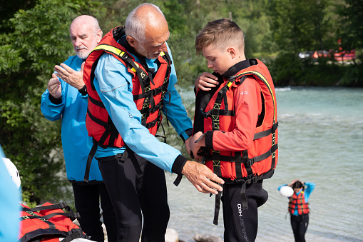 Senior man helping boy how to correctly fasten life jacket before the rafting.